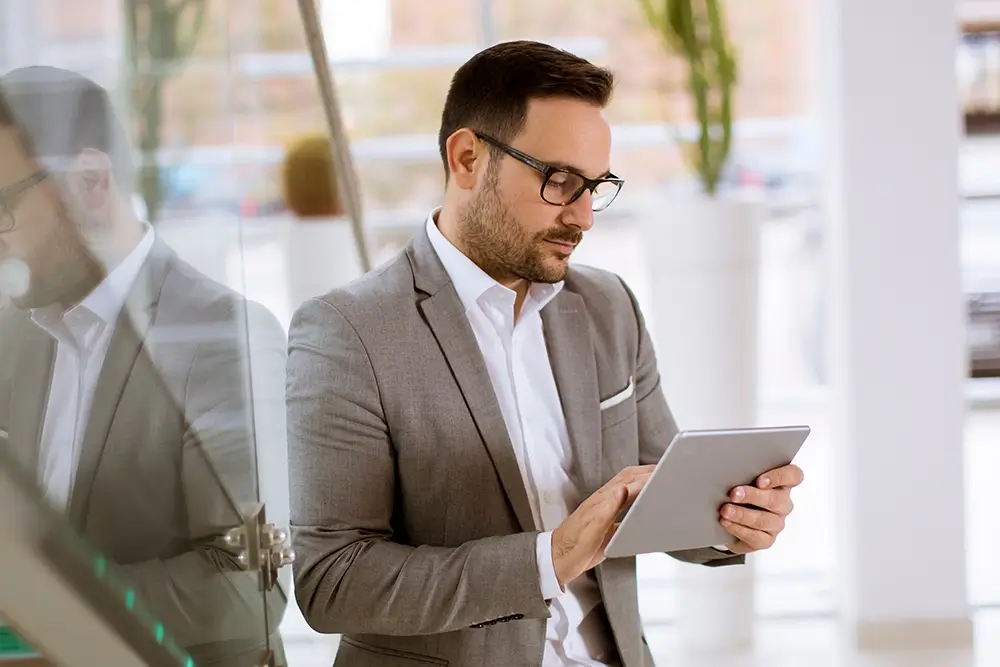 A professional property manager in a grey suit and glasses using a digital tablet for transparent fire safety communication. He is standing in a brightly lit, modern office lobby, representing the integration of technology in building management.