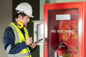 A professional safety inspector wearing a white hard hat, high-visibility yellow vest, and ear protection using a digital tablet and stylus to perform a fire safety equipment check. He is documenting the condition of a red fire hose cabinet in a commercial facility.