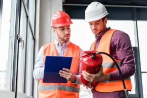 Two professional safety inspectors in hard hats and high-visibility vests conducting a fire safety audit. One inspector holds a clipboard while the other examines a red fire extinguisher near a large window in a modern commercial building.