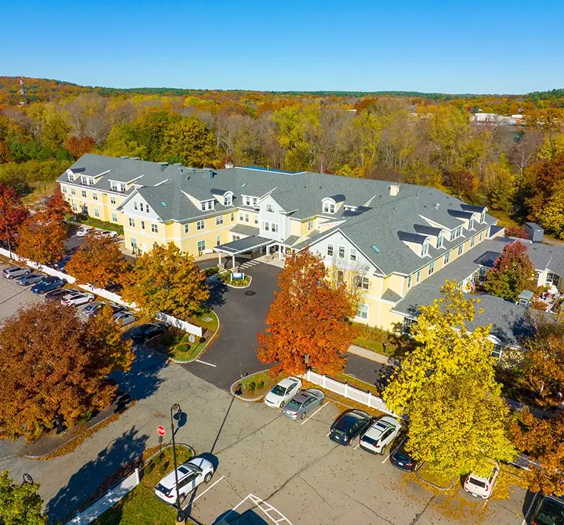 Aerial view of a sprawling yellow senior living community building with a grey roof, surrounded by colorful autumn trees and a paved parking lot.