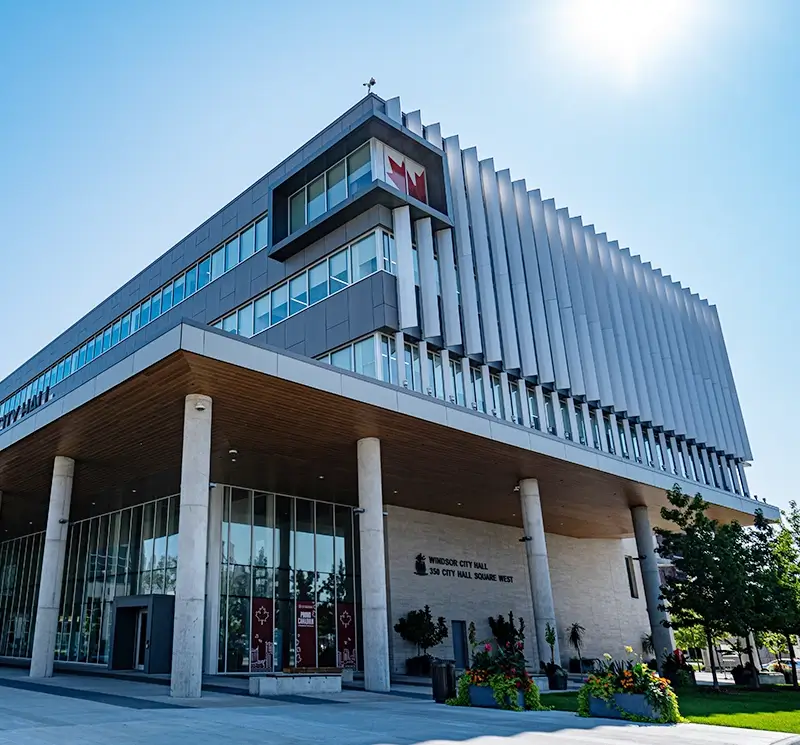 Modern architectural view of Windsor City Hall featuring a cantilevered roof with wood paneling, large glass windows, and vertical metal fins under a bright sun.