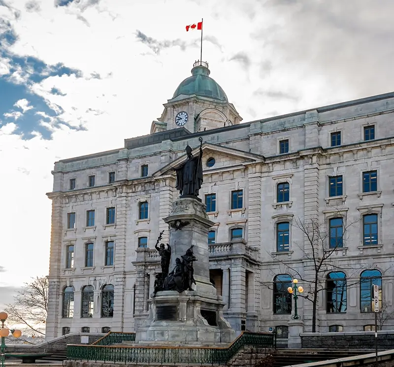 Historic stone government building with a green copper dome, a clock tower flying the Canadian flag, and a large bronze monument in the foreground.