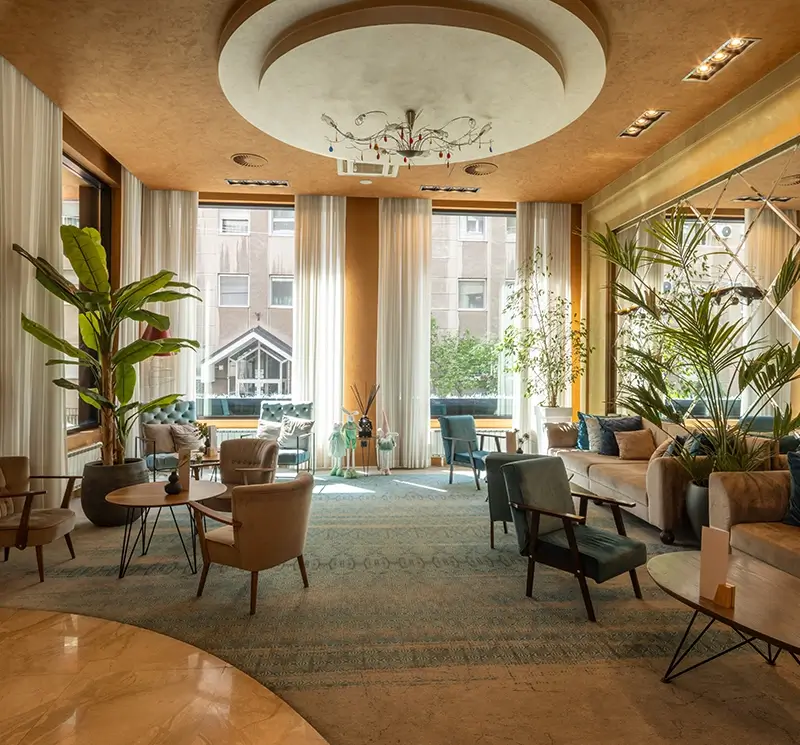 Modern hotel lounge with mid-century style armchairs, indoor potted plants, and large floor-to-ceiling windows with white curtains.