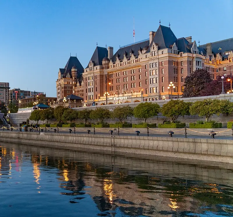 Historic grand hotel with chateau-style architecture reflecting in the calm water of a harbor during twilight.