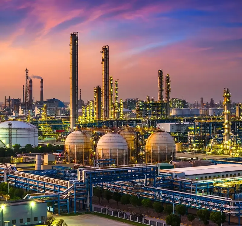 A large industrial refinery complex with tall distillation towers, spherical storage tanks, and extensive piping networks under a vibrant sunset sky.