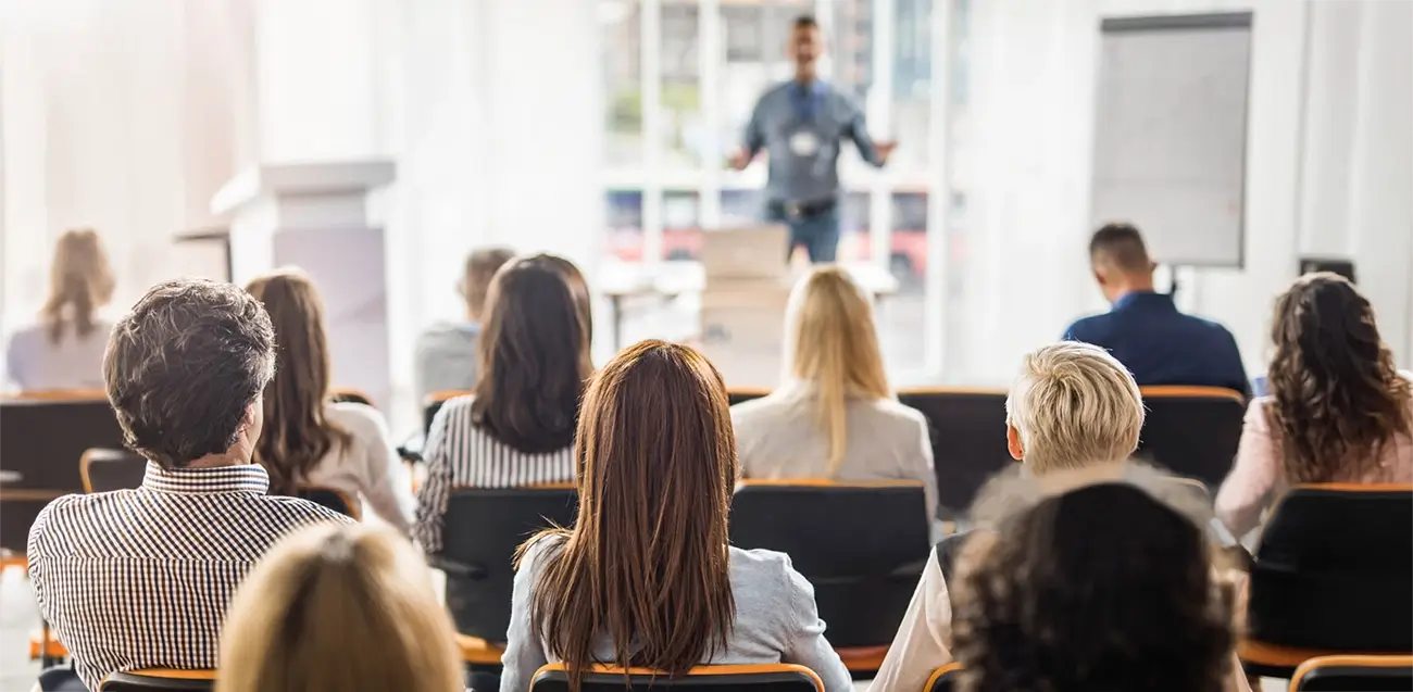 Instructor-led emergency training session for property staff in a seminar room setting. On-Demand vs Instructor-Led Emergency Training - A view from the back of a brightly lit seminar room where a group of people is seated in rows, facing a speaker at the front. The instructor, seen slightly out of focus in the distance, stands near a podium and a whiteboard while delivering a presentation or training session.