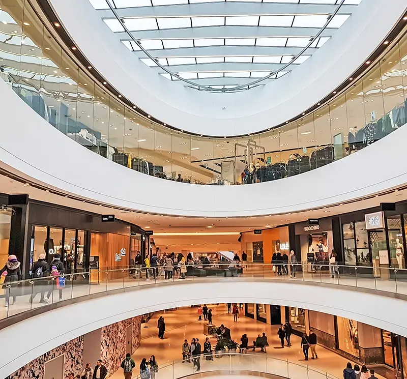 Interior view of a multi-level modern shopping mall with a large curved glass skylight and shoppers walking past retail stores.
