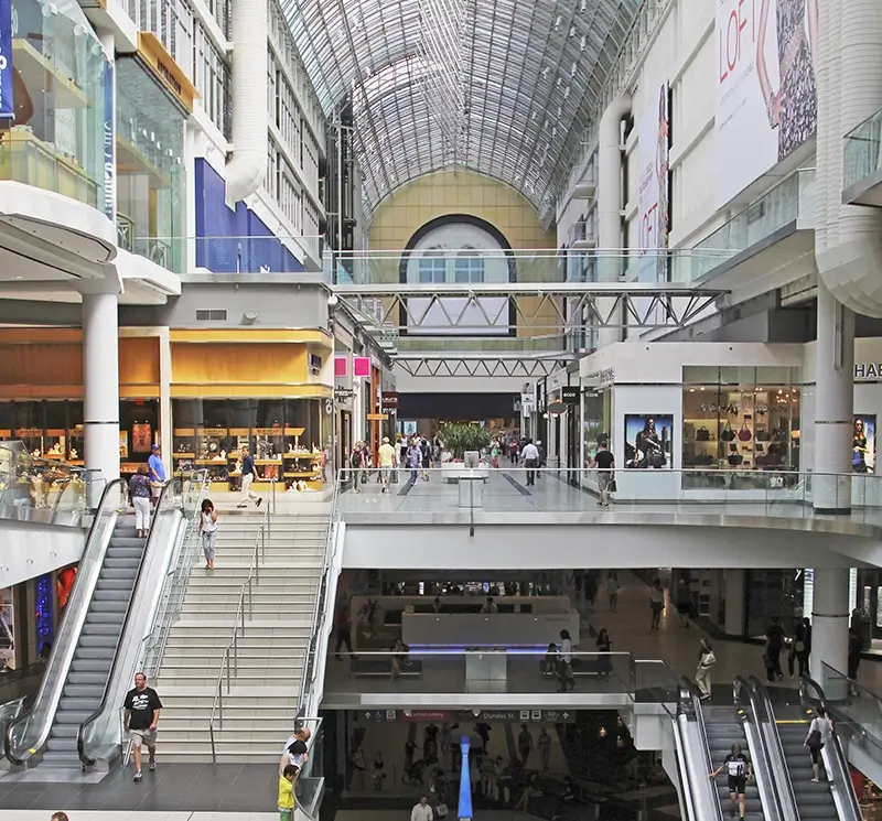 Bright interior of a multi-story urban shopping center featuring escalators, pedestrian bridges, and a high arched glass ceiling.